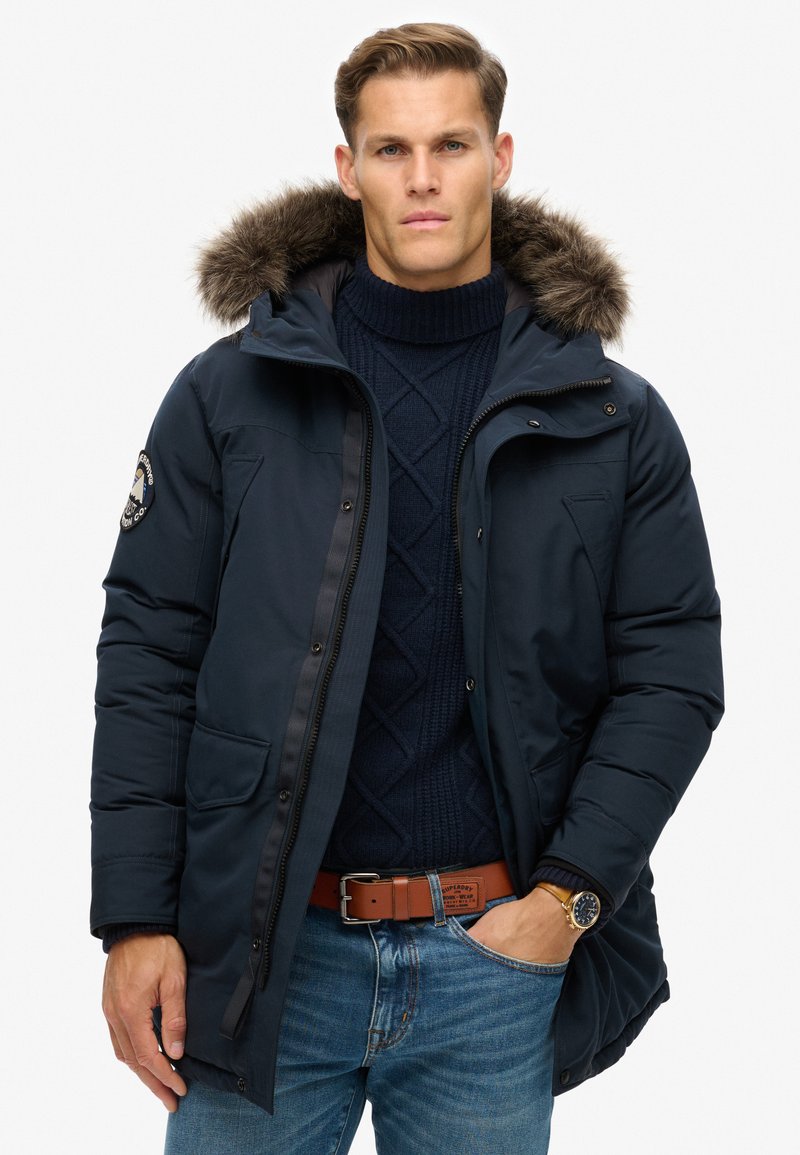 Marineblauer Parkajacke mit Kunstfellkragen, zwei Fronttaschen und Reißverschluss. Getragen über einem marineblauen Strickpullover und hellblauen Jeans.