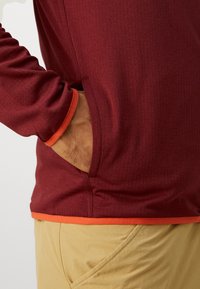 Mano en el bolsillo lateral de una camisa de manga larga roja con detalles naranjas, combinada con pantalones beige.