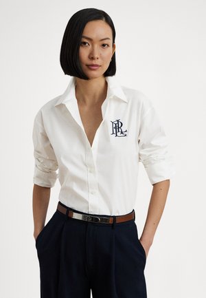 Camicia bianca con bottoni e scollo a V profondo, maniche arrotolate e logo ricamato blu scuro sul petto a sinistra. Indossata con pantaloni scuri e cintura.