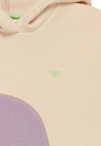 Felpa beige con una forma lavanda sul davanti, con un piccolo logo a cuore verde sul petto. Tessuto morbido con una superficie liscia.