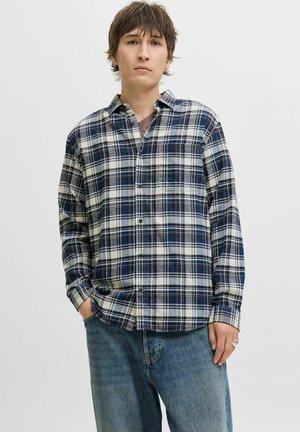 Camisa de cuadros en tonos azul, blanco y oscuros; de mangas largas, con cuello abotonado, bolsillo en el pecho, corte relajado, tejido texturizado. Llevada con vaqueros azules.