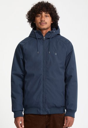 Mann trägt eine marineblaue Kapuzenjacke mit Frontreißverschluss und Kordeln, steht vor einem schlichten weißen Hintergrund.