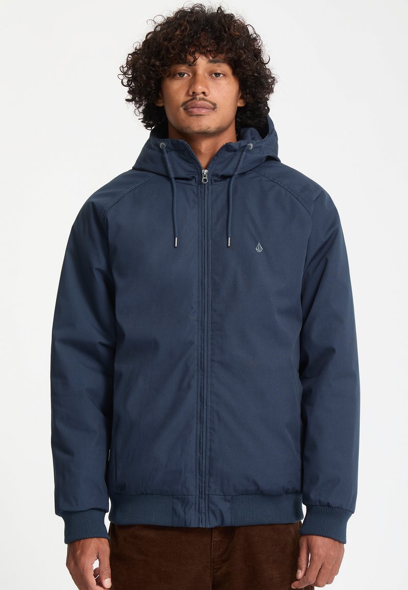 Homme portant une veste à capuche bleu marine avec une fermeture éclair à l’avant et des cordons, debout devant un fond blanc uni.
