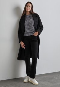 Schwarzer Mantel über einem strukturierten grauen Pullover, kombiniert mit schwarzen Jeans und weißen Sneakers. Glatter Stoff, taillierte Passform und neutrale Farbpalette.