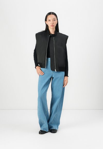 Gilet imbottito nero con spalle larghe, cerniera argento, indossato sopra un top nero. I jeans svasati blu chiaro e le scarpe nere completano l'outfit.