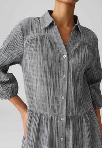 Grijze gestreepte shirtdress met een knoopsluiting aan de voorkant, kraag en opbollende mouwen. Gemaakt van lichte stof met een geraffineerde taille en zakken.