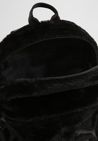 Sac à dos en peluche noir avec un extérieur texturé, une fermeture éclair et un intérieur doux. Équipe d'un petit logo en relief à l'avant.