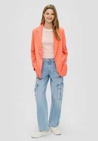 Perzikkleurige blazer, witte en rode gestreepte top, lichtblauwe cargobroek en witte sneakers. De blazer heeft een getailleerd ontwerp en grote zakken.