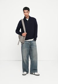 Marineblauer Zip-Up-Pullover, hellgraue Umhängetasche, weit geschnittene, ausgewaschene Jeans und sportliche weiße und graue Schuhe. Minimalistisches Design.