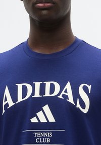 Μπλε βαμβακερό t-shirt με λευκό λογότυπο "ADIDAS", γραφικό σε σχήμα τριγώνου και κείμενο "TENNIS CLUB". Κλασικός σχεδιασμός με στρογγυλή λαιμόκοψη. Ομαλή υφή.