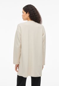 Μπεζ μακρυμάνικη ζακέτα με άνετη εφαρμογή, υφή υφάσματος, πέτα ώμων και ευθεία γραμμή στο τελείωμα, φορεμένη πάνω από μαύρο παντελόνι.
