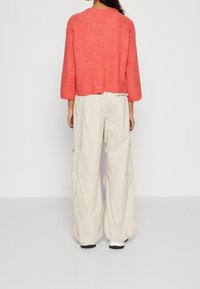 Korallinv ärinen neulottu villapaita, jossa leveät hihat, yhdistettynä kermaan vivahtaviin cargo-housuihin, joissa on sivutaskut, yksinkertaista valkoista taustaa vasten.