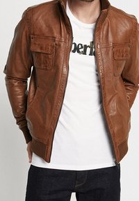 Giacca di pelle marrone con zip anteriore, orlo e polsini a coste, due tasche sul petto e una texture liscia, indossata sopra una camicia bianca.