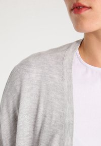 Ljusgrå cardigan med en mjuk, stickad textur, öppen frontdesign och ribbade kanter, lagd över en vit skjorta.