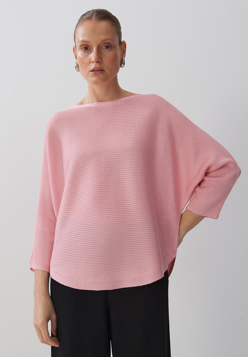 Haut en tricot côtelé rose clair avec un large col et des manches dolman, associé à un pantalon noir. Texture douce, coupe décontractée et ourlet arrondi.