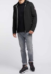 Veste noire zippée avec capuche et accents gris, associée à un jean gris et des baskets noires aux semelles blanches, illustrant une tenue décontractée.