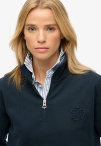 Sweat à zippé marine en tissu texturé, avec un monogramme subtil, porté sur une chemise à col rayée bleu clair.
