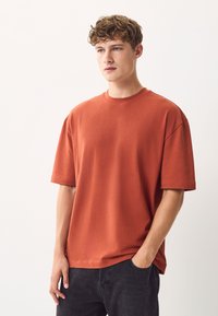 Rozsdaszínű, puha anyagú, oversized póló kerek nyakkivágással és rövid ujjakkal, sötét farmernadrággal párosítva, minimalista dizájn.