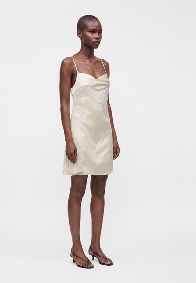 Femme portant une robe nuisette beige en satin longueur genou avec des détails en dentelle et des bretelles fines, associée à des sandales noires à talons.