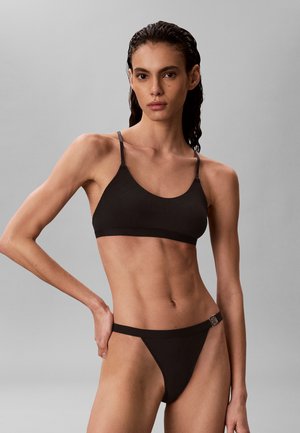 Femme aux cheveux mouillés portant un soutien-gorge noir et un bas de bikini taille haute, debout devant un fond gris uni.