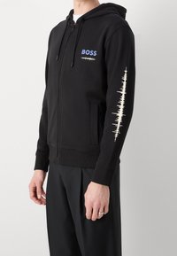 Felpa nera con zip in cotone misto. Presenta il logo "BOSS" in blu, con un grafico ad onda sonora sul manica sinistra. Tasche frontali incluse.