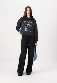 Un hoodie noir avec un imprimé graphique coloré et des étoiles, associé à un jean noir à jambes larges. Le modèle tient un sac à bandoulière bleu clair.
