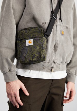 Personne portant un sweat à capuche Carhartt gris délavé, un pantalon cargo foncé et un sac bandoulière Carhartt vert à motif paisley avec une sangle noire.