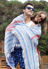Large striped beach blanket in blue and white with red accents, fringed edges. Two people wrapped in it, outdoors with greenery in background.