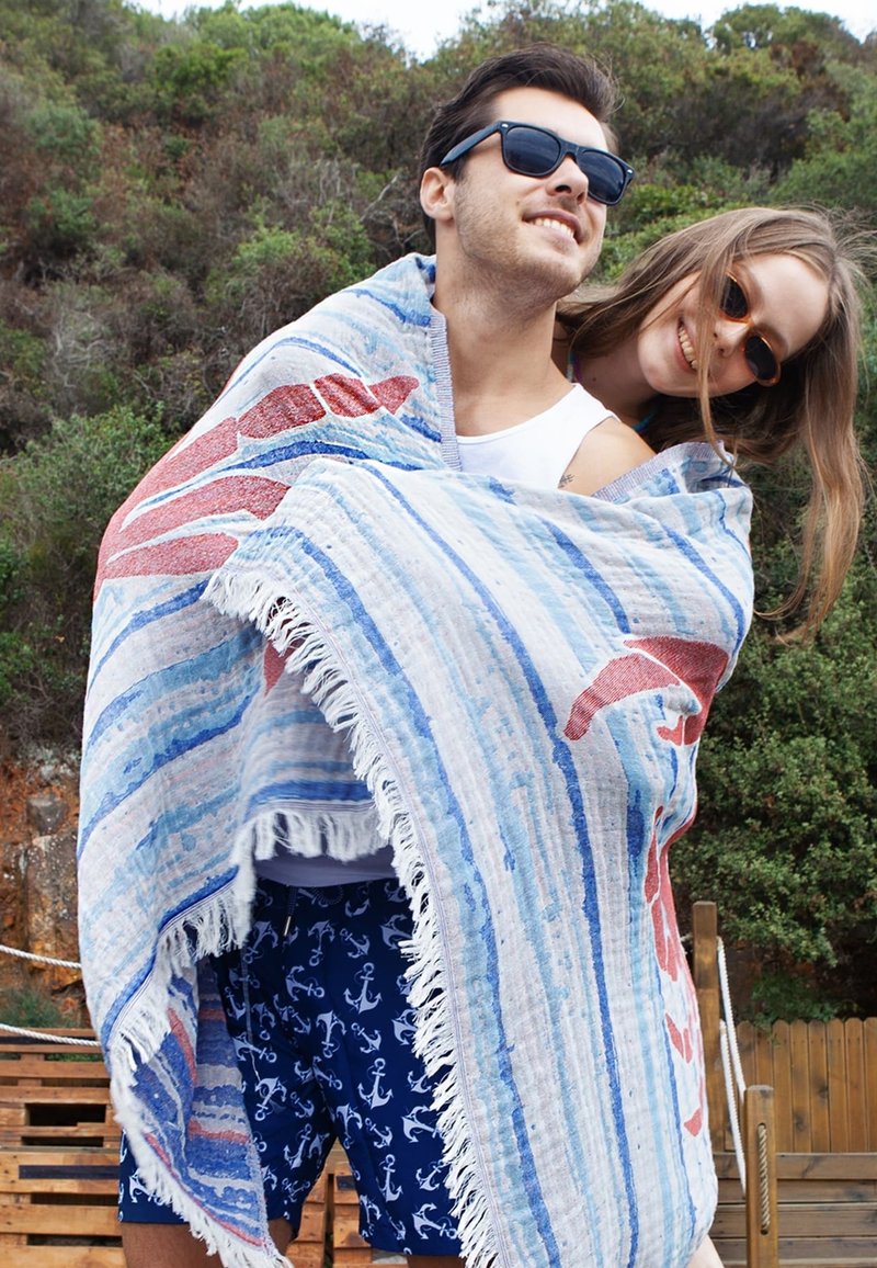 Large striped beach blanket in blue and white with red accents, fringed edges. Two people wrapped in it, outdoors with greenery in background.