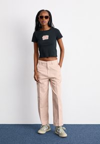 Svart cropped t-skjorte med "Santa Cruz" grafikk, beige rette bukser og grønne sneakers. Tilbehør inkluderer brune solbriller.