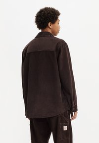 Brown corduroy shirt with a relaxed fit, featuring a shirt collar, long sleeves, and a chest pocket with a label at the hem.