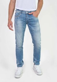 Pantalones vaqueros de mezclilla azul claro con corte slim, que presentan bolsillos frontales, una textura desgastada y costuras en contraste, combinados con zapatillas blancas.