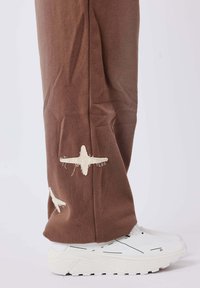 Pantaloni marroni con accenti di cucitura bianca sfilacciata ai lati, abbinati a sneaker bianche spesse con suole testurizzate.