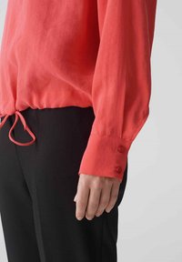 Blusa corallo con maniche lunghe, cordoncino regolabile all'orlo e polsini con bottone, abbinata a pantaloni neri. Tessuto liscio e leggero.