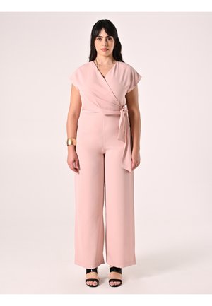 Roze wikkeljumpsuit met korte mouwen, een strikdetail in de taille en broekspijpen met wijde pijpen. Gecombineerd met zwarte sandalen met bandjes.