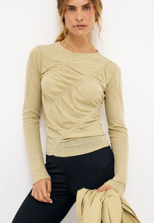 Žena s blond vlasmi má na sebe béžový dlhý rukáv s riasením, čierne nohavice a drží zladenú béžovú bundu na bielom pozadí.