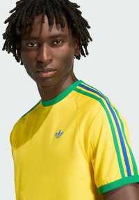 Młody mężczyzna z krótkimi dredami, ubrany w żółtą koszulkę Adidas z zielonymi i niebieskimi paskami na ramionach, na jasnym tle.