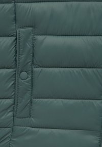 Tissu matelassé vert foncé avec un détail de patte de boutonnage à pressions verticales sur une veste rembourrée.