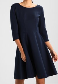 Granatowa sukienka w kształcie litery A z rękawami o długości trzy czwarte, okrągłym dekoltem i plisowanym dołem. Gładka tekstura materiału, bez widocznych wzorów czy elementów metalowych.