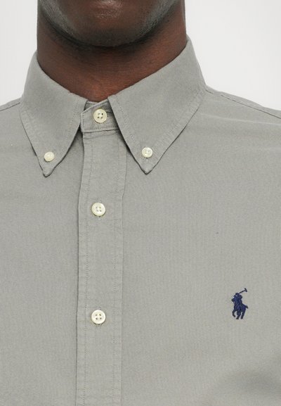 Siva srajca s gumbi, izdelana iz strukturiranega blaga, z klasičnim ovratnikom, belimi gumbi in majhnim modrim izvezenim logotipom na prsih.