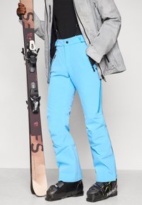 Mėlynos slidinėjimo kelnės su užtrauktukais šoninėse dalyse, pagamintos iš vandeniui atsparios medžiagos, derinamos su juodais slidinėjimo batais ir tekstūruotu pilku slidinėjimo švarku.