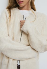 Cárdigan de punto en crema con textura acanalada, mangas largas y cierre delantero con cremallera. La tela parece suave y ligeramente holgada para un ajuste cómodo.