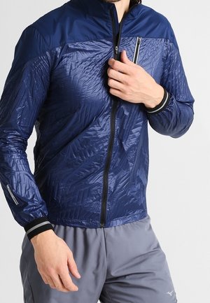 Mężczyzna noszący błyszczącą granatową kurtkę na zamek z wzorzystą teksturą oraz szare spodnie, regulujący zamek kurtki.