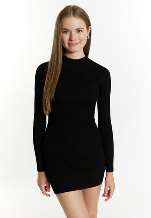 Junge Frau mit langem glattem Haar, die ein figurbetontes schwarzes Mini-Kleid mit langen Ärmeln und blaue runde Ohrringe trägt, lächelt in die Kamera.