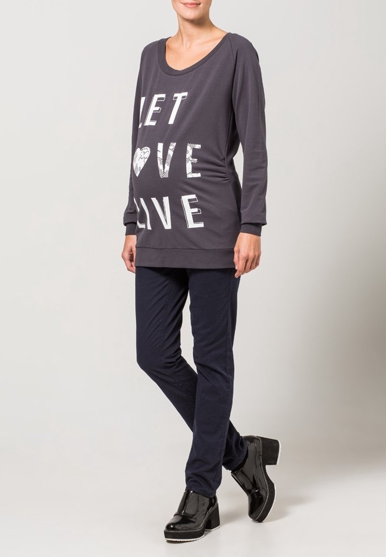 Femme portant un haut à manches longues foncé avec le texte "LET LOVE LIVE" et un motif cœur, pantalon slim foncé et chaussures à plateforme noires, debout.