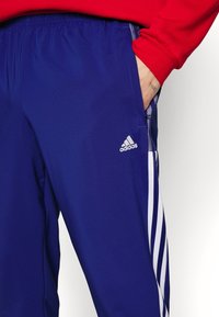 Niebieskie spodnie sportowe z białym logo i paskami Adidas, z elastycznym pasem w talii oraz teksturowanym panelem bocznym. Kieszeń widoczna z boku.