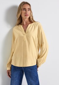 Blusa a maniche lunghe con scollo a V in morbido tessuto rayon giallo chiaro. Presenta polsini elastici e una vestibilità comoda. Abbinata a jeans in denim blu.