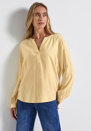 Blouse à manches longues et décolleté en v en doux tissu de rayonne jaune pâle. Présente des poignets élastiqués et une coupe décontractée. Associée à un jean en denim bleu.