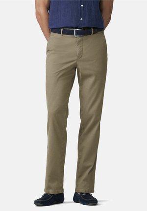 Mann trägt beige Chinos, navy-gewebten Gürtel, navy Loafers und ein in die Hose gestecktes dunkelblaues Hemd mit Knöpfen, steht mit den Händen in den Taschen.