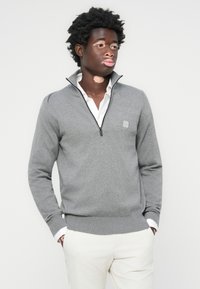 Grau zipfeliger Pullover aus weichem Material mit Stehkragen, gerippten Bündchen und Saum, kombiniert mit einem weißen Hemd und hellen Hosen.
